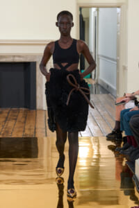 Model on a runway wearing a black crop top and a textured, ruffled black skirt tied with a brown rope belt; gold pointed heels gleam as she walks.