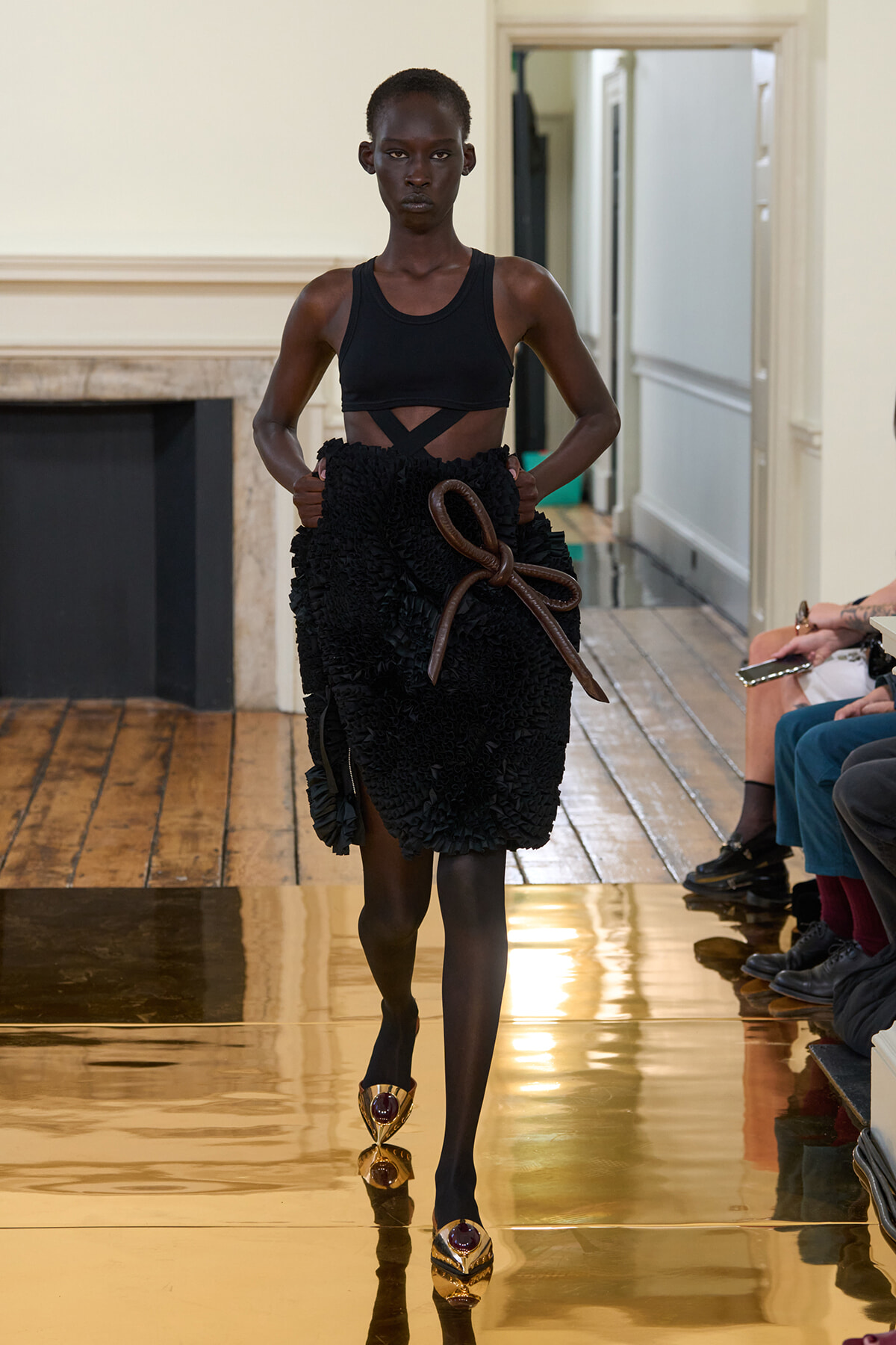 Model on a runway wearing a black crop top and a textured, ruffled black skirt tied with a brown rope belt; gold pointed heels gleam as she walks.