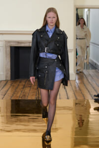 Model walking a runway in a black leather oversized jacket with a belt over a blue shirt-dress; gold-pointed toe shoes visible on glossy floor.