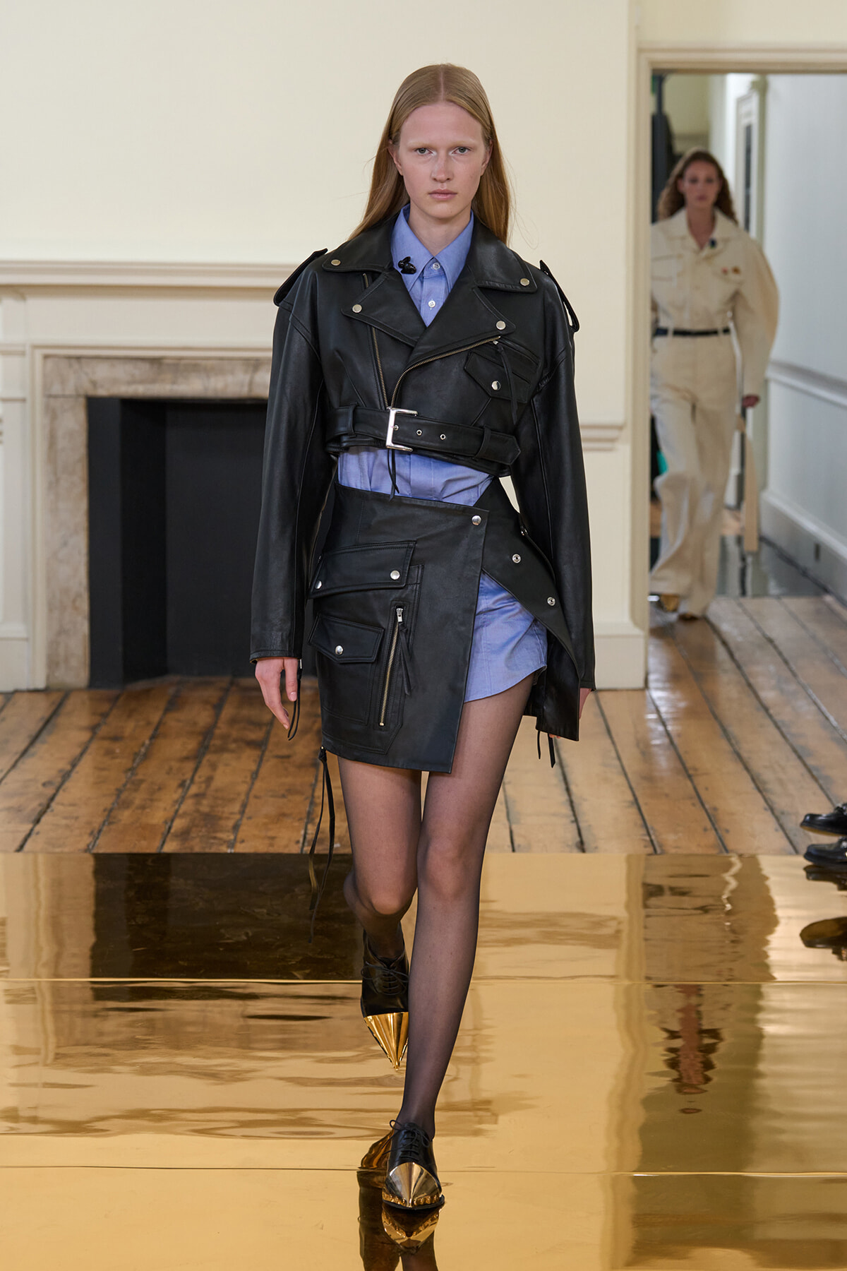 Model walking a runway in a black leather oversized jacket with a belt over a blue shirt-dress; gold-pointed toe shoes visible on glossy floor.