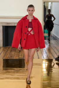 Model walking a runway in a bold red oversized coat over a matching red crop top and mini skirt.