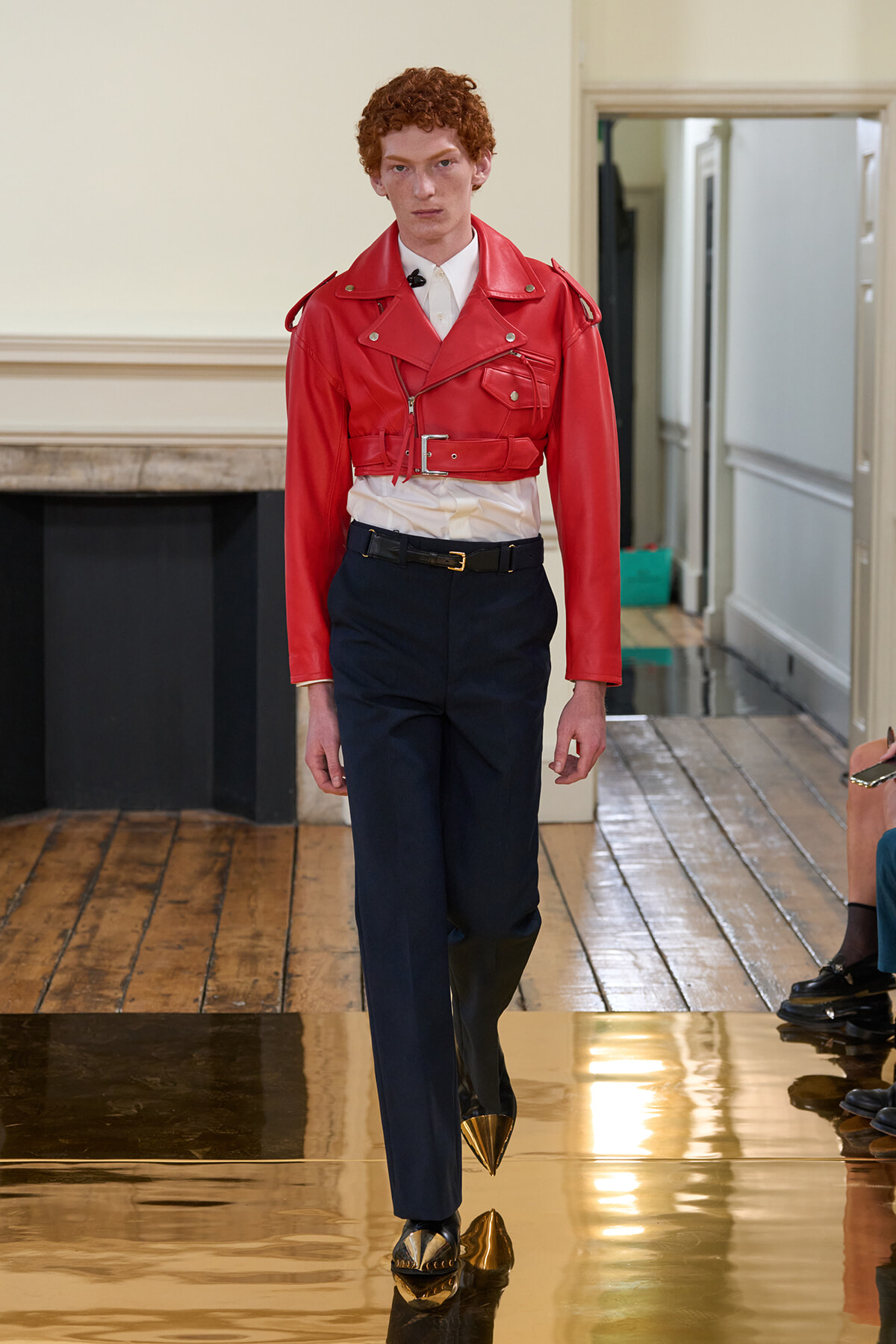 Model wearing a red cropped leather jacket over a white shirt with navy trousers and gold pointed shoes on a fashion runway.