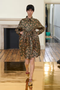 Model walking a runway in a dark floral print dress with a cinched waist and red strappy heels on a gold-tinted glossy floor.