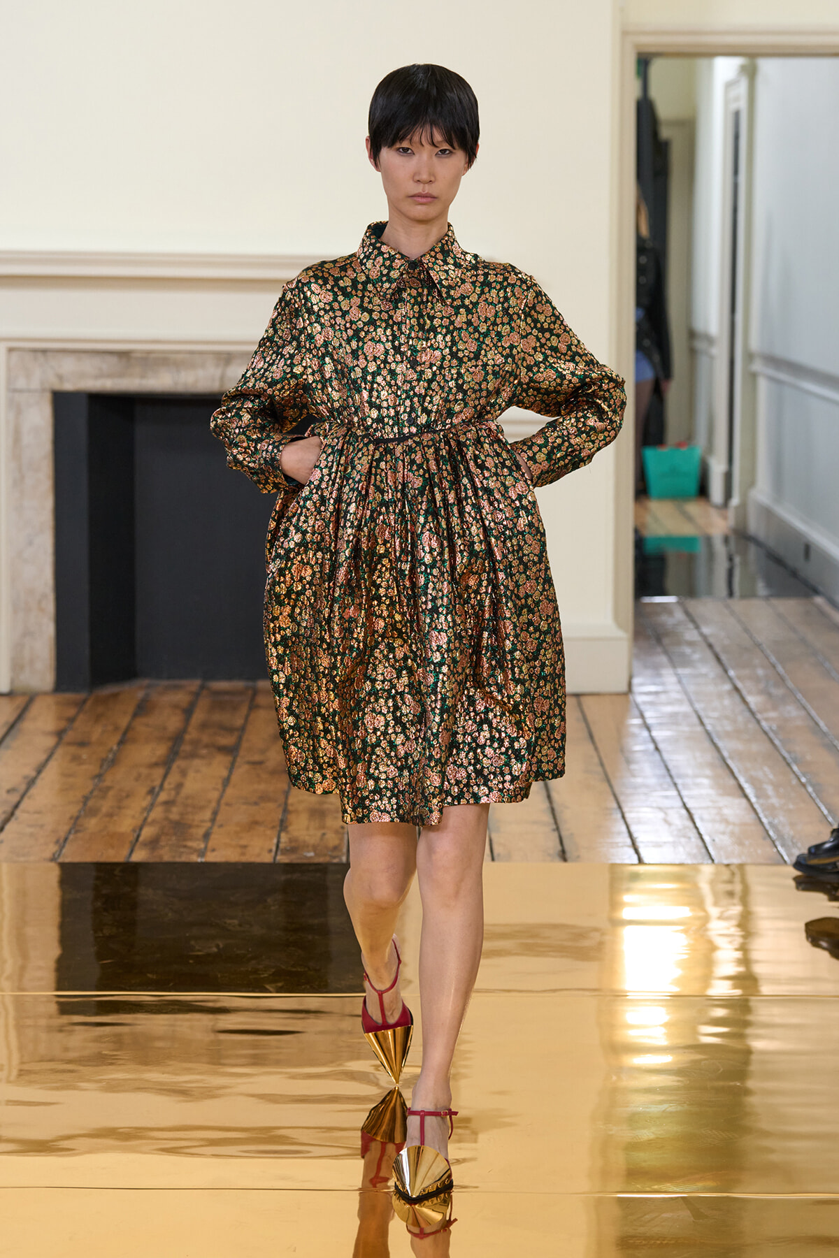 Model walking a runway in a dark floral print dress with a cinched waist and red strappy heels on a gold-tinted glossy floor.