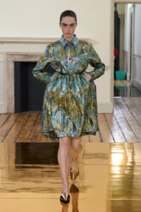 Model walks a gold runway wearing a metallic green, blue, and gold belted dress with long sleeves with hands in pockets.