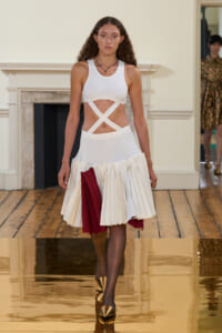 Model walking a fashion runway in a white crop top with crisscross waist straps and a white pleated skirt with burgundy panels, wearing gold pointed-toe shoes.