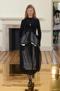 Model walking a runway in a black fitted top with a layered sheer gray and black skirt, blue beaded necklace, and gold shoes with green stones.