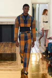 Male model on a fashion runway wearing a bold plaid patchwork suit in mustard, brown, and navy, with a dark top and metallic gold shoes.