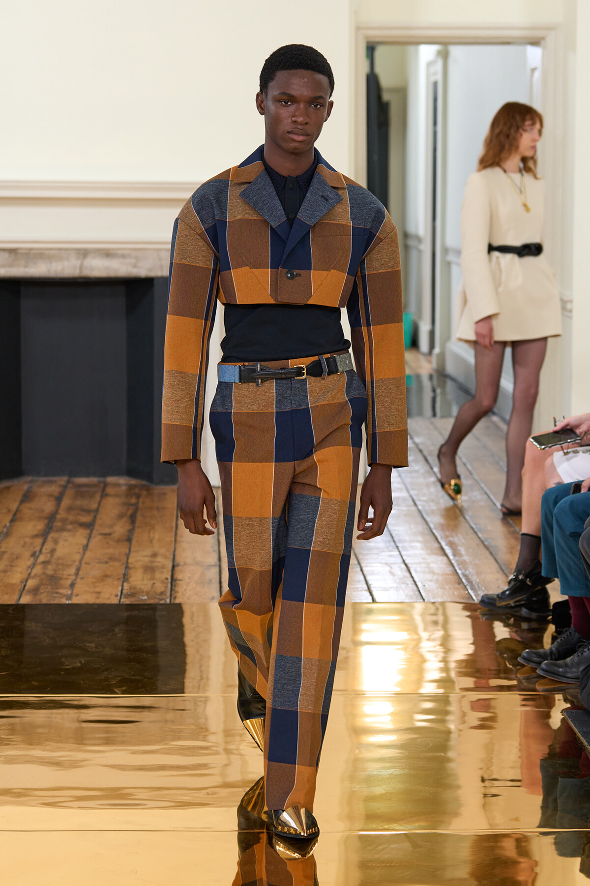 Male model on a fashion runway wearing a bold plaid patchwork suit in mustard, brown, and navy, with a dark top and metallic gold shoes.