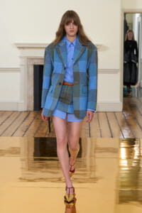 Model walks a fashion runway in a blue plaid oversized blazer over a light blue shirt and matching shorts, with a brown belt and metallic heels.