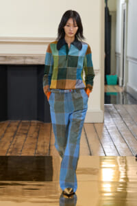 Model wearing a color-block patchwork blouse with a black collar and matching blue plaid pants, walking on a runway indoors.