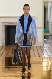 Model walks a fashion runway in a light blue shirt under a transparent plastic coat with black lapels, blue shorts, and black tights, carrying a clear tote and wearing gold-toe heels on a glossy floor.