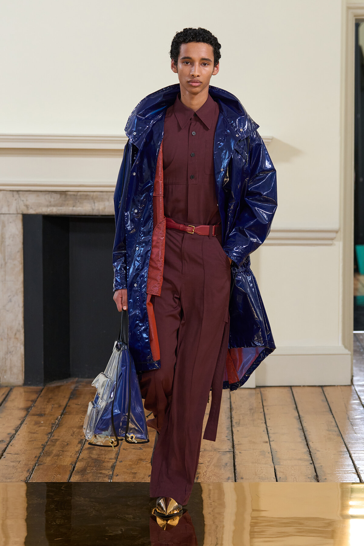 Model walks a runway in a burgundy suit with a shiny navy raincoat, holding a clear blue handbag.