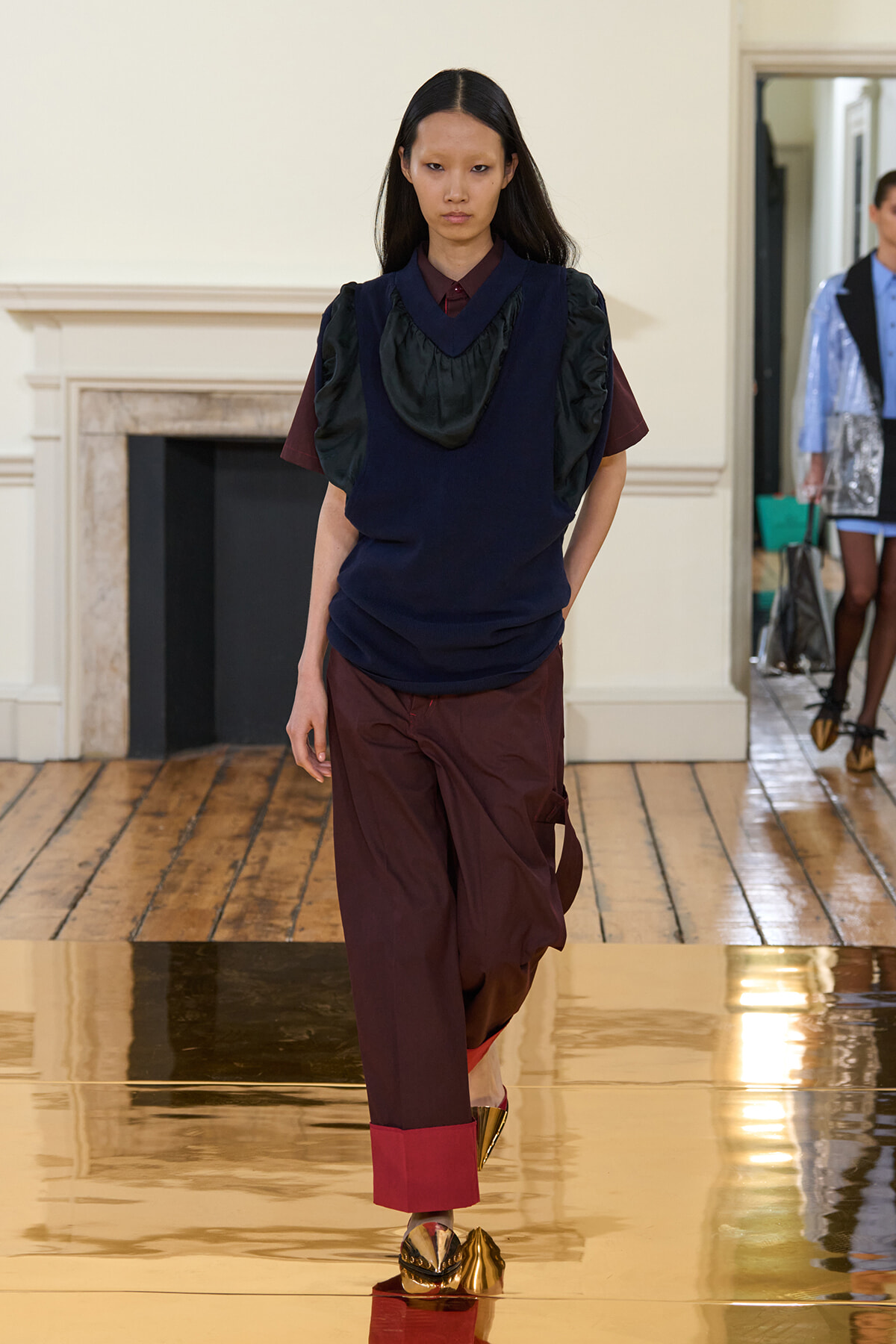 Model walks a fashion-show runway indoors in a navy layered top over burgundy pants, with bold gold shoes.