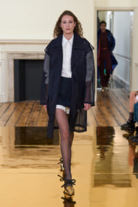 Model walks a fashion show in a sheer navy coat over a white shirt and navy shorts, gold bow-tie heels reflecting on the glossy floor.