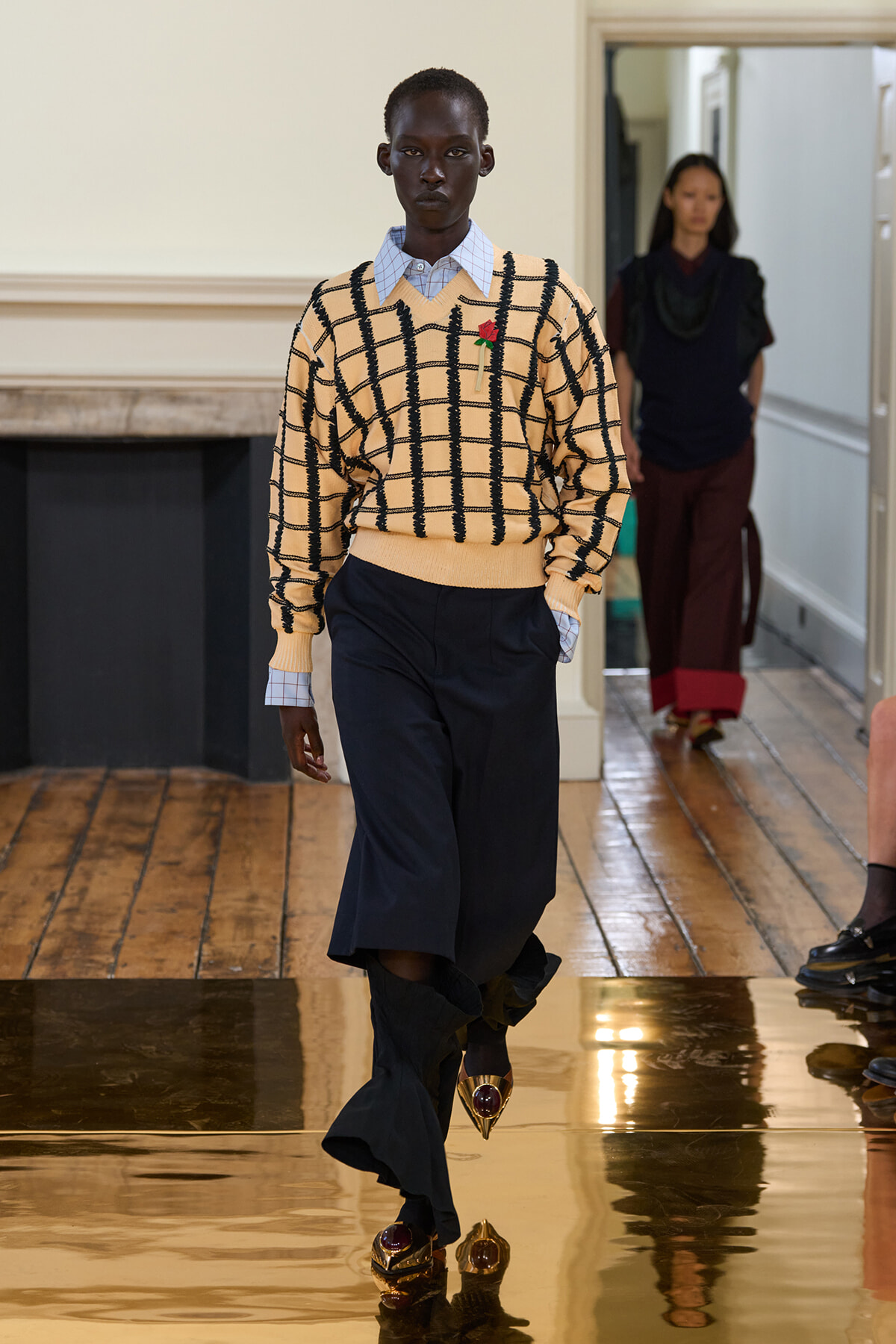Model walks a glossy runway in a beige grid-knit sweater over a white collared shirt, navy wide-leg pants, and jeweled gold shoes.