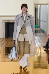 Model walking a runway in a color-block beige and gray trench over a tan suit, carrying a clear bag on a gold-reflective floor.