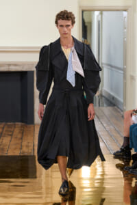 Male model on a runway wearing an oversized black coat with a voluminous skirt-like lower half and belt; black-and-gold shoes