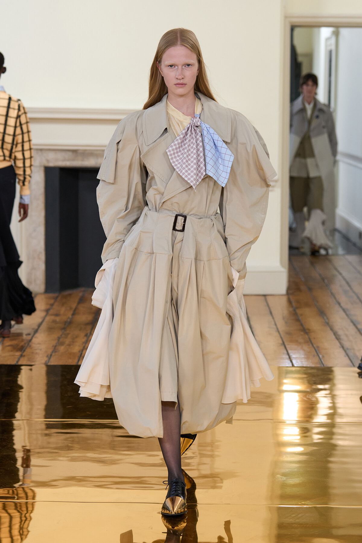 Model walking a fashion runway in a voluminous beige trench-coat dress with a tied scarf and black-and-gold oxford shoes.
