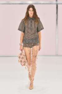 Model on runway in a checkered short-sleeve shirt and sheer pink embroidered pants, carrying a pink handbag.