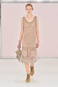 Model walking a runway in a sheer beige lace dress with a boxy hem and tan boots, carrying a patterned bag.