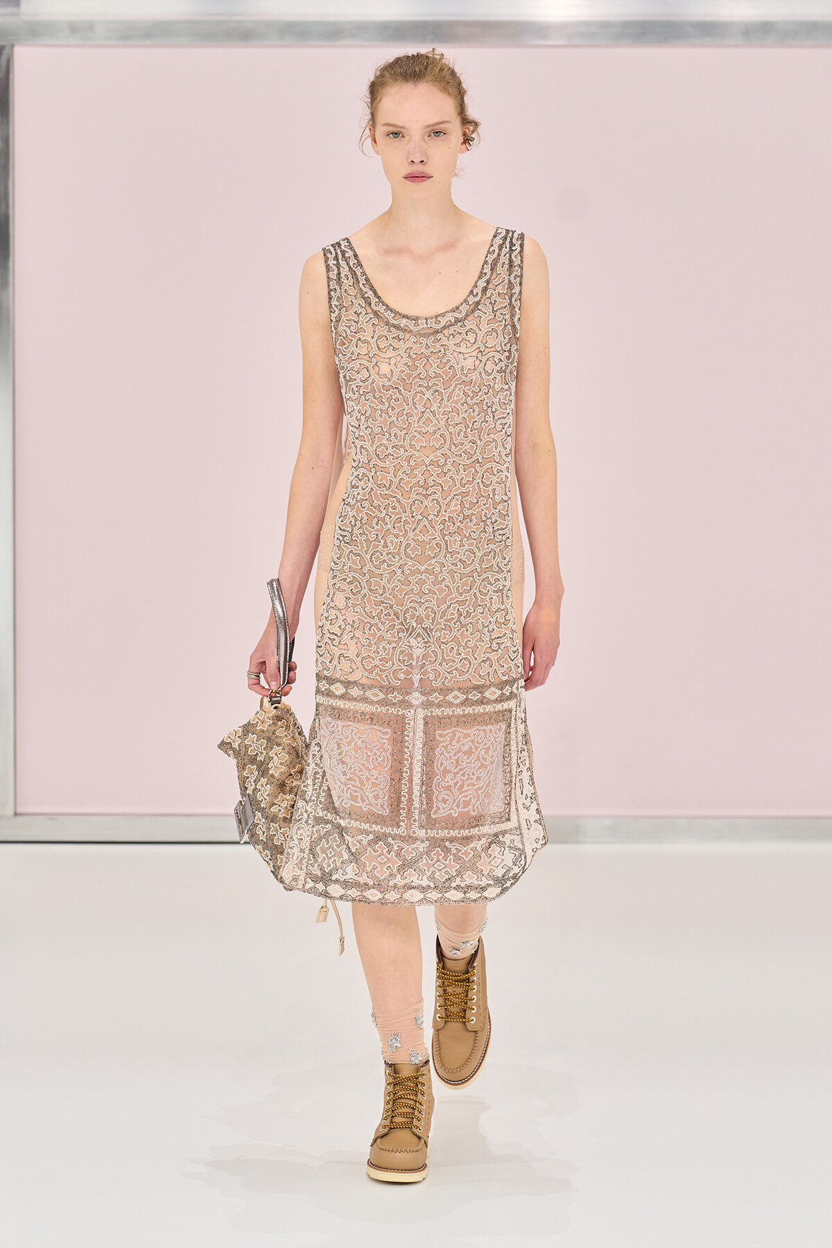 Model walking a runway in a sheer beige lace dress with a boxy hem and tan boots, carrying a patterned bag.