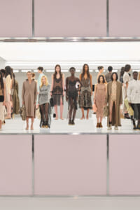 Group of models standing on white risers in a fashion show, wearing neutral-toned dresses and coats against a pale pink backdrop.