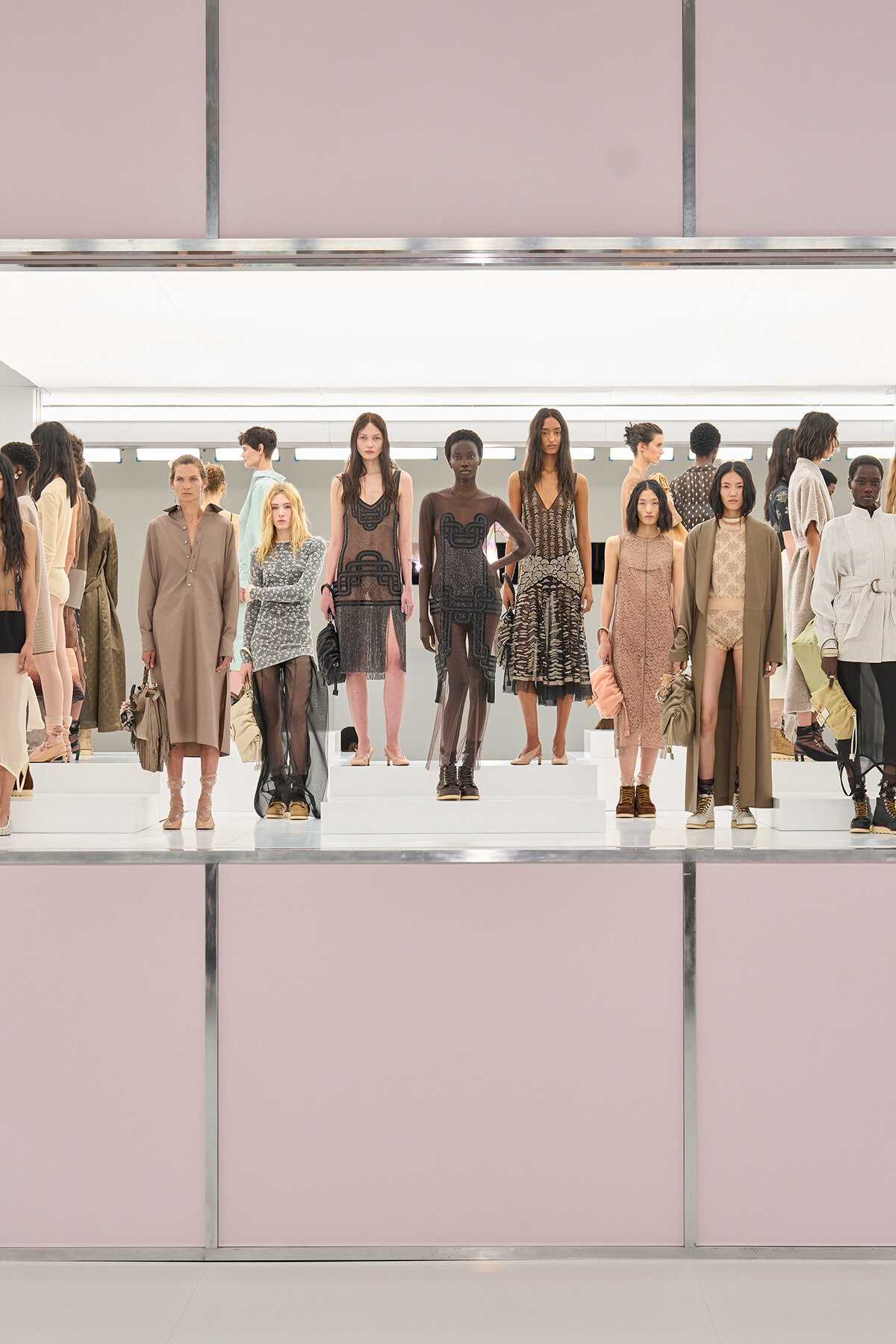 Group of models standing on white risers in a fashion show, wearing neutral-toned dresses and coats against a pale pink backdrop.