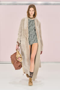 Model on a runway wearing a long beige faux-fur coat over a green floral romper, tan lace-up boots, and a large brown handbag with a pink tassel.