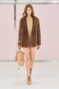 Model walking runway in a textures brown coat over a beige cardigan, carrying a tan handbag; blush pink boots and socks visible.