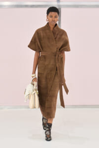 Model walking a fashion runway in a brown knit wrap dress with a waist tie, carrying a light-colored handbag and wearing embellished black boots.