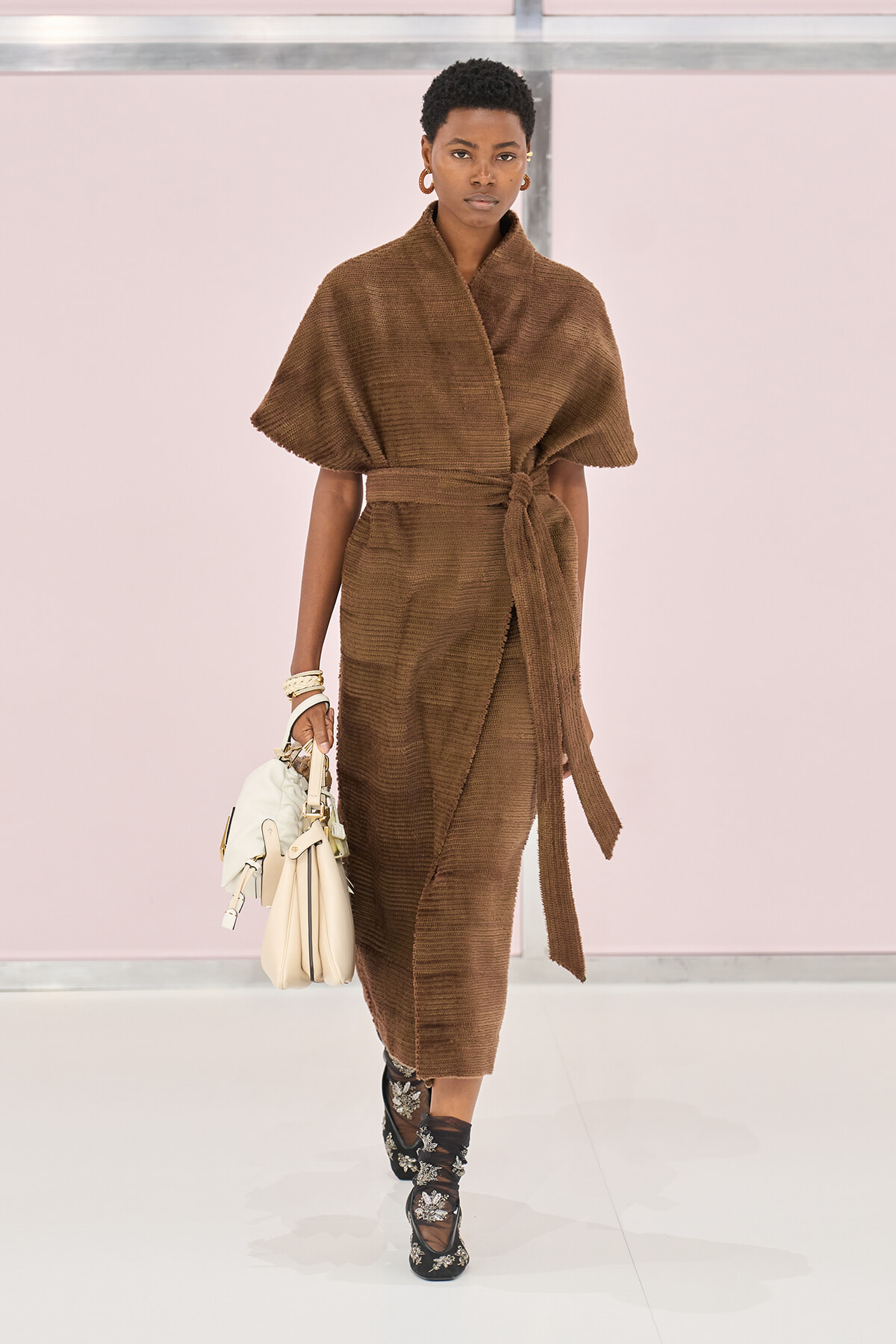 Model walking a fashion runway in a brown knit wrap dress with a waist tie, carrying a light-colored handbag and wearing embellished black boots.