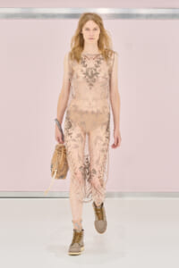 Model walking a runway in a sheer pale pink jumpsuit with metallic floral embroidery, carrying a tan handbag and tan boots on the catwalk.
