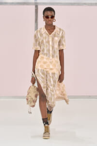 Model walking a runway in a cream and olive floral print shirt and matching skirt with a front slit, accessorized with brown sunglasses, hoop earrings, and a textured cream bag.