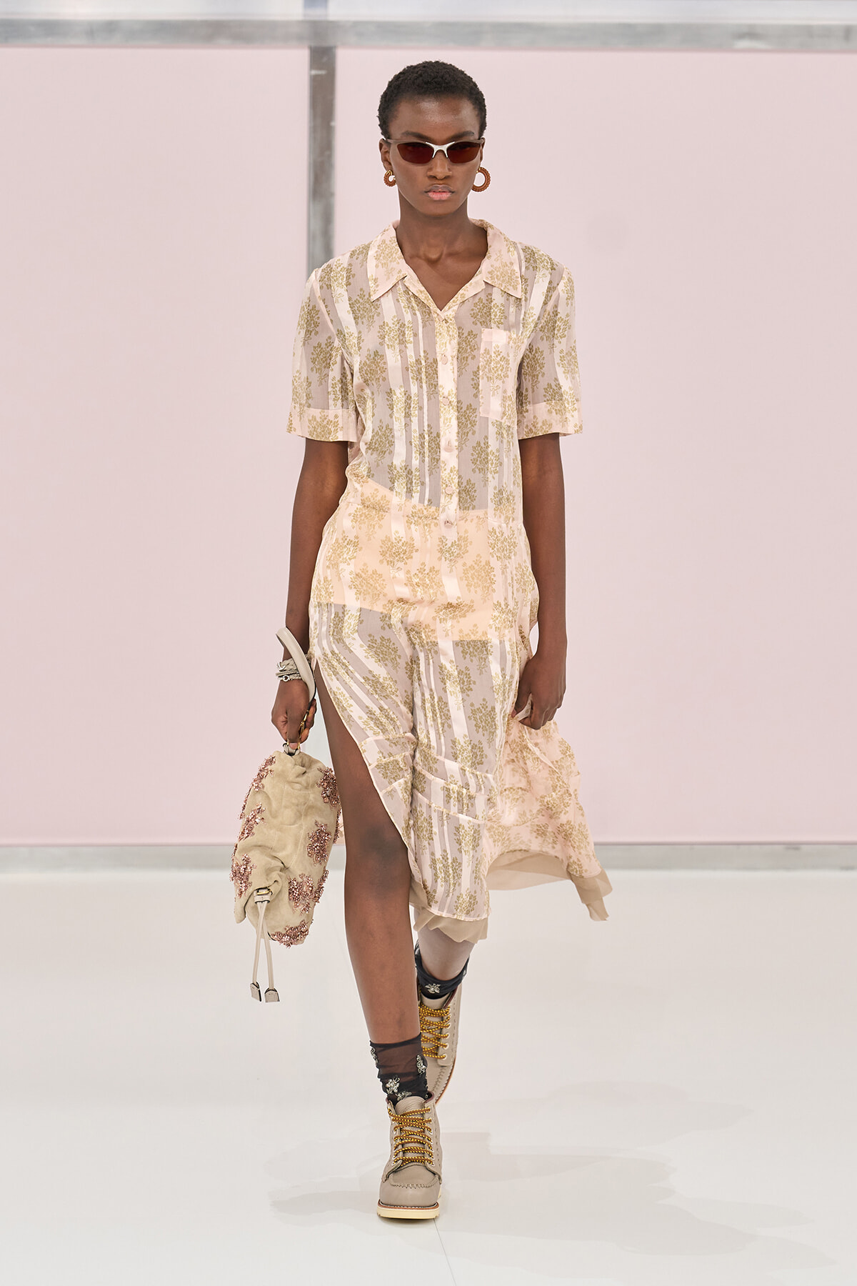 Model walking a runway in a cream and olive floral print shirt and matching skirt with a front slit, accessorized with brown sunglasses, hoop earrings, and a textured cream bag.