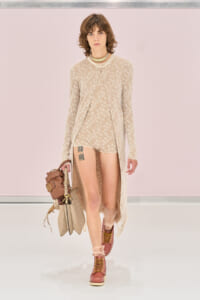 Model walking the runway in a beige floral knit set (cardigan and shorts) with pink lace-up boots and tan handbags in hand.