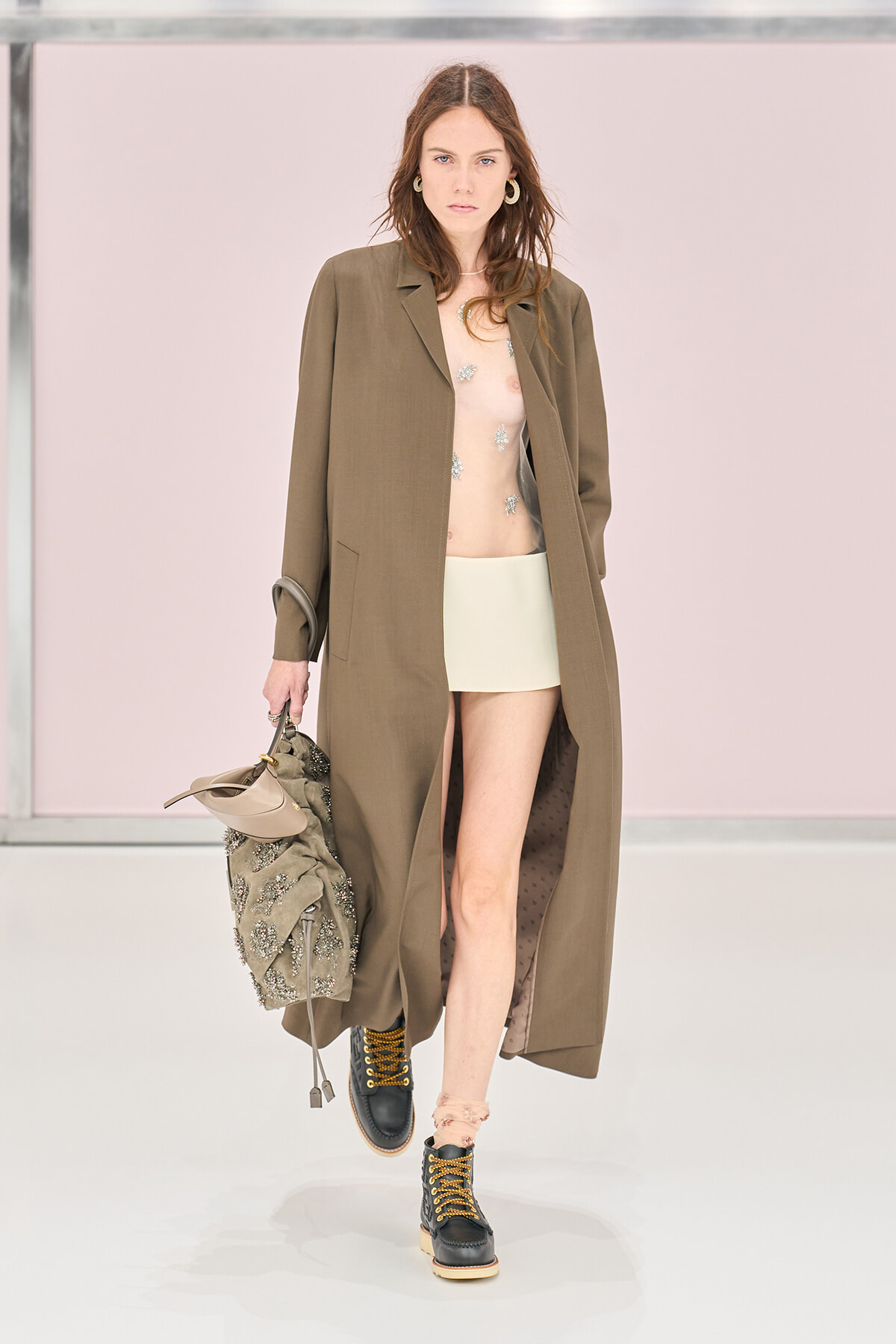 Model walking a runway in a long brown coat over a sheer, sequined top and pale mini skirt, paired with black lace-up boots and a beaded bag.