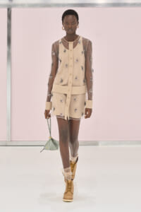 Model walking the runway in a beige sheer-sleeve mini dress with floral embroidery, tan lace-up boots, and a pale green handbag.