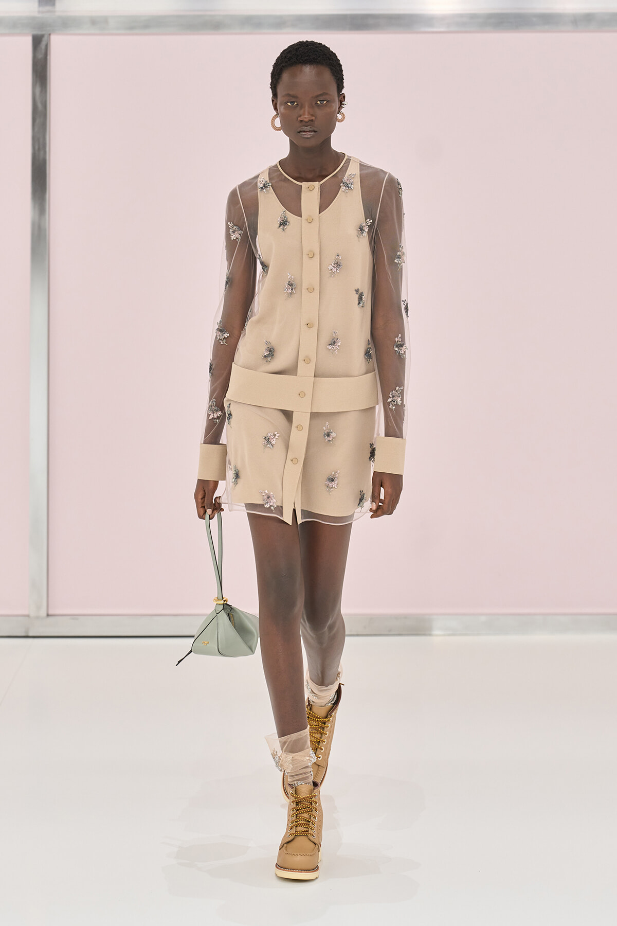 Model walking the runway in a beige sheer-sleeve mini dress with floral embroidery, tan lace-up boots, and a pale green handbag.