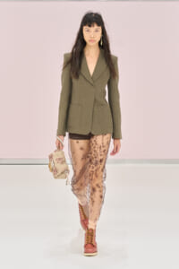 Model on a runway wearing an olive blazer with oversized shoulders and large front pockets, paired with a sheer pink embroidered skirt, pink boots, and a beige embellished handbag.