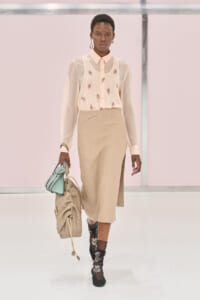 Model walks a runway in a pale beige blouse with embroidery and a tan midi skirt, holding a beige drawstring bag and a mint-green purse.