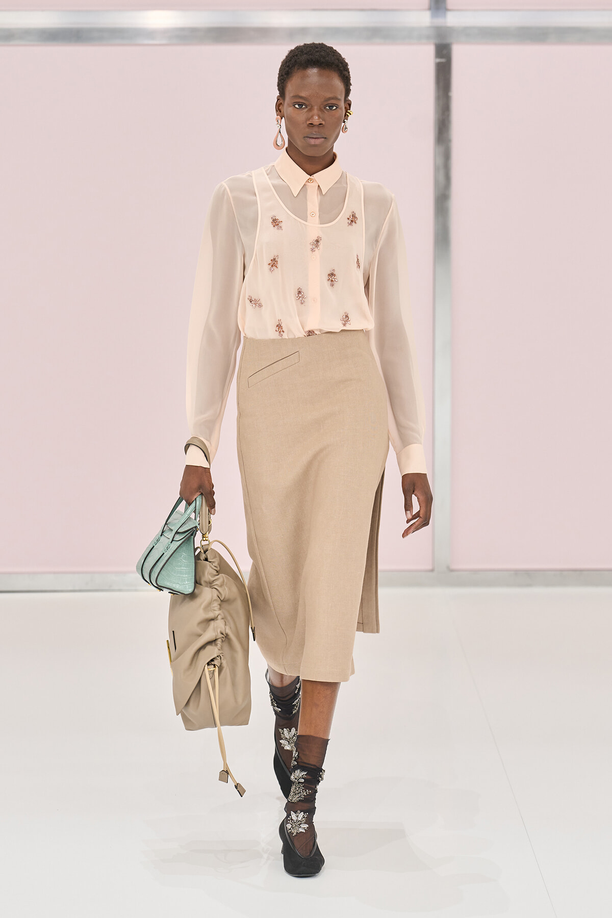 Model walks a runway in a pale beige blouse with embroidery and a tan midi skirt, holding a beige drawstring bag and a mint-green purse.