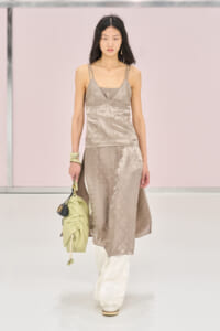 Model walking a runway in a taupe satin slip top and matching midi skirt, white pants, carrying a pale green handbag and wearing bracelets.
