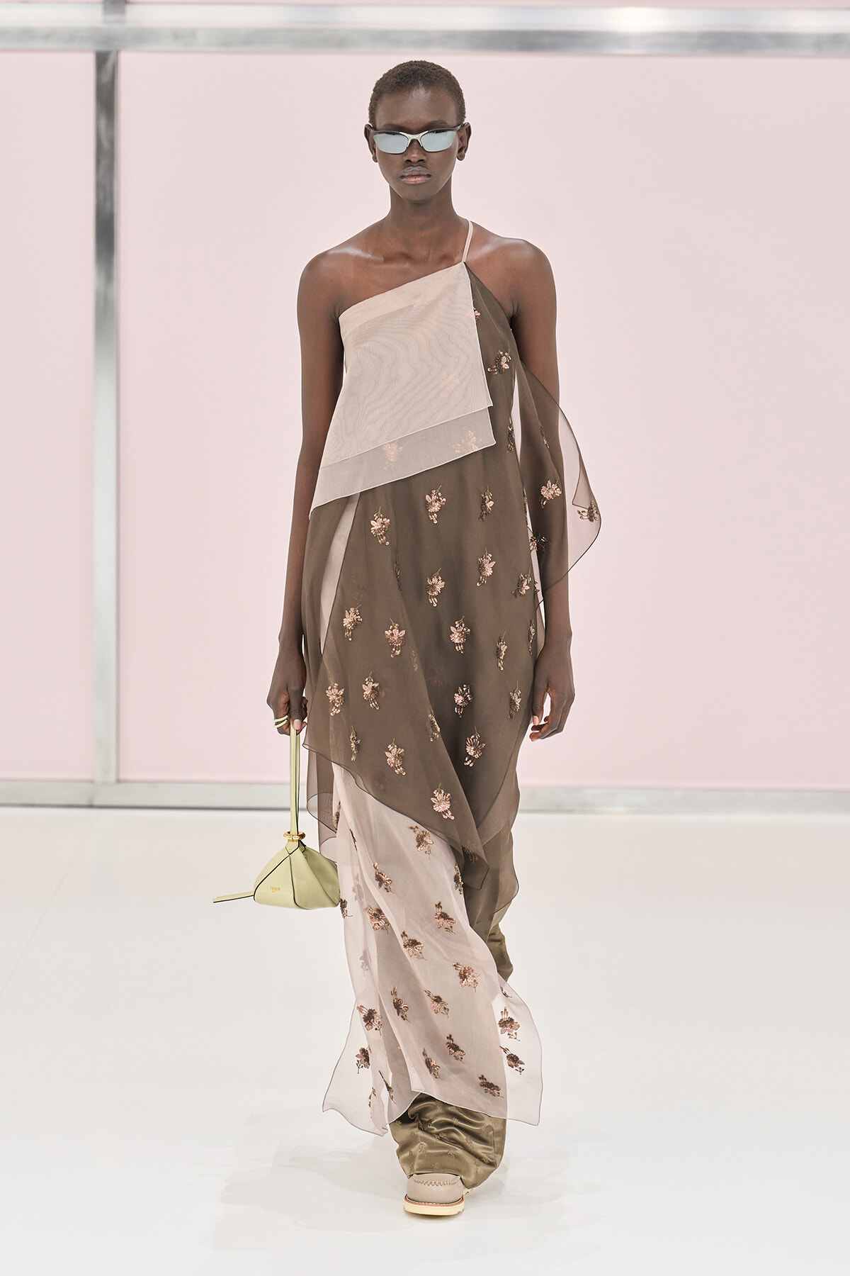 Model on a runway wearing a sheer, embroidered, asymmetrical dress with layered panels, sunglasses, and a small pale yellow bag.