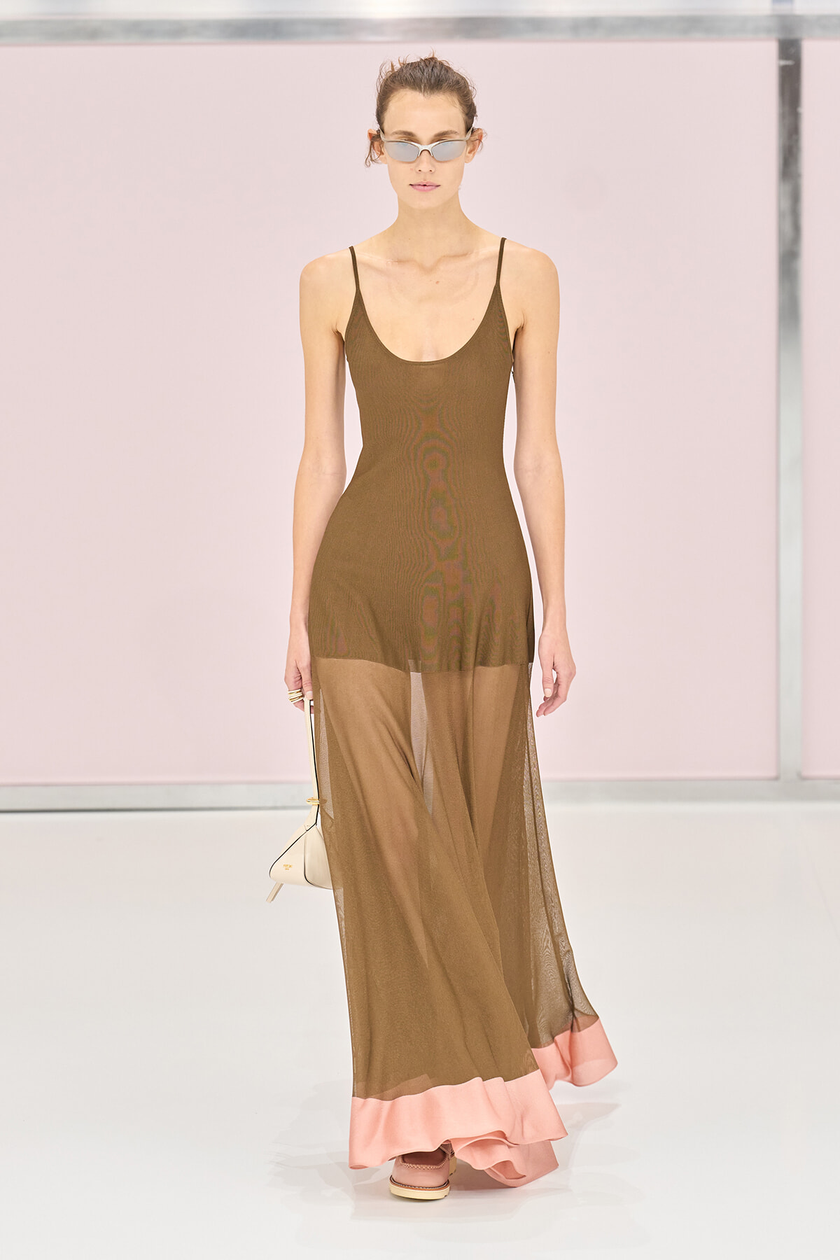 Model walking a runway in a brown spaghetti-strap sheer maxi dress with a pink ruffle hem, wearing sunglasses and carrying a small white handbag.