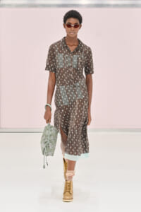 Runway model in a sheer brown midi dress with white floral print, mint panels, and pale blue trim; carries a mint embroidered bag and wears sunglasses and tan boots.