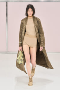 Model walking a runway in a beige knit outfit with an olive long coat, carrying a patterned bag and wearing tan boots.