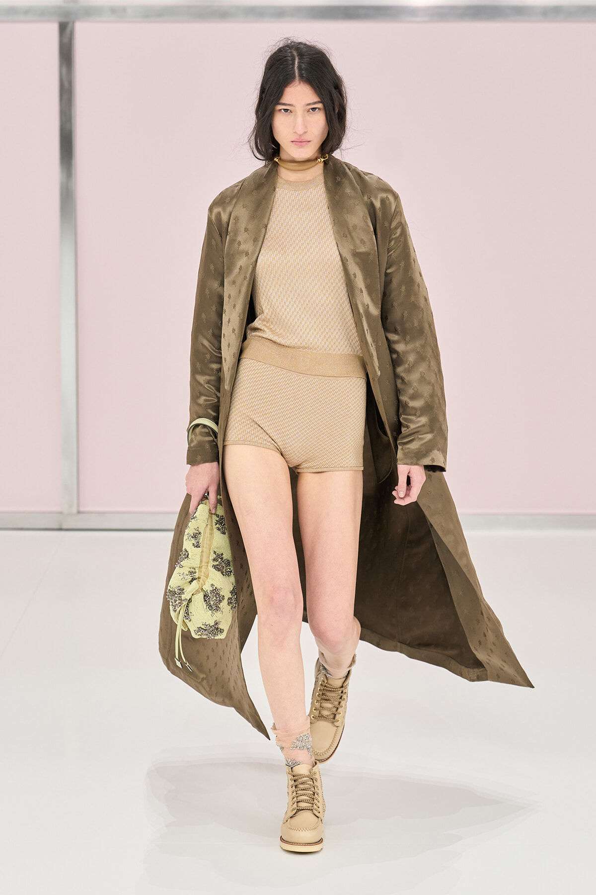 Model walking a runway in a beige knit outfit with an olive long coat, carrying a patterned bag and wearing tan boots.
