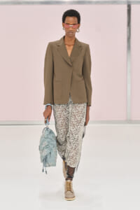 Model walking the runway in a brown tailored blazer with large patch pockets, white lace pants, tan lace-up boots, pink sunglasses, and gold hoops, carrying a light blue embroidered bag.