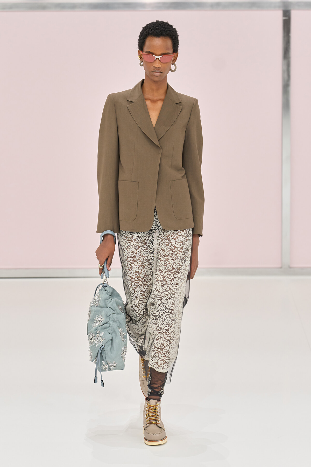 Model walking the runway in a brown tailored blazer with large patch pockets, white lace pants, tan lace-up boots, pink sunglasses, and gold hoops, carrying a light blue embroidered bag.